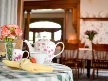 A tea pot and cup rests on a checkered cloth table with fresh pastries and carnations.